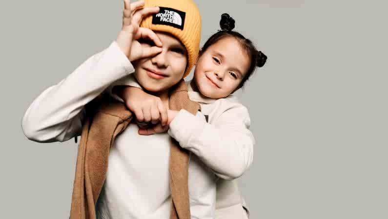 Двое детей в уютной одежде позируют на сером фоне. Девочка обнимает мальчика, носящего жёлтую шапку.