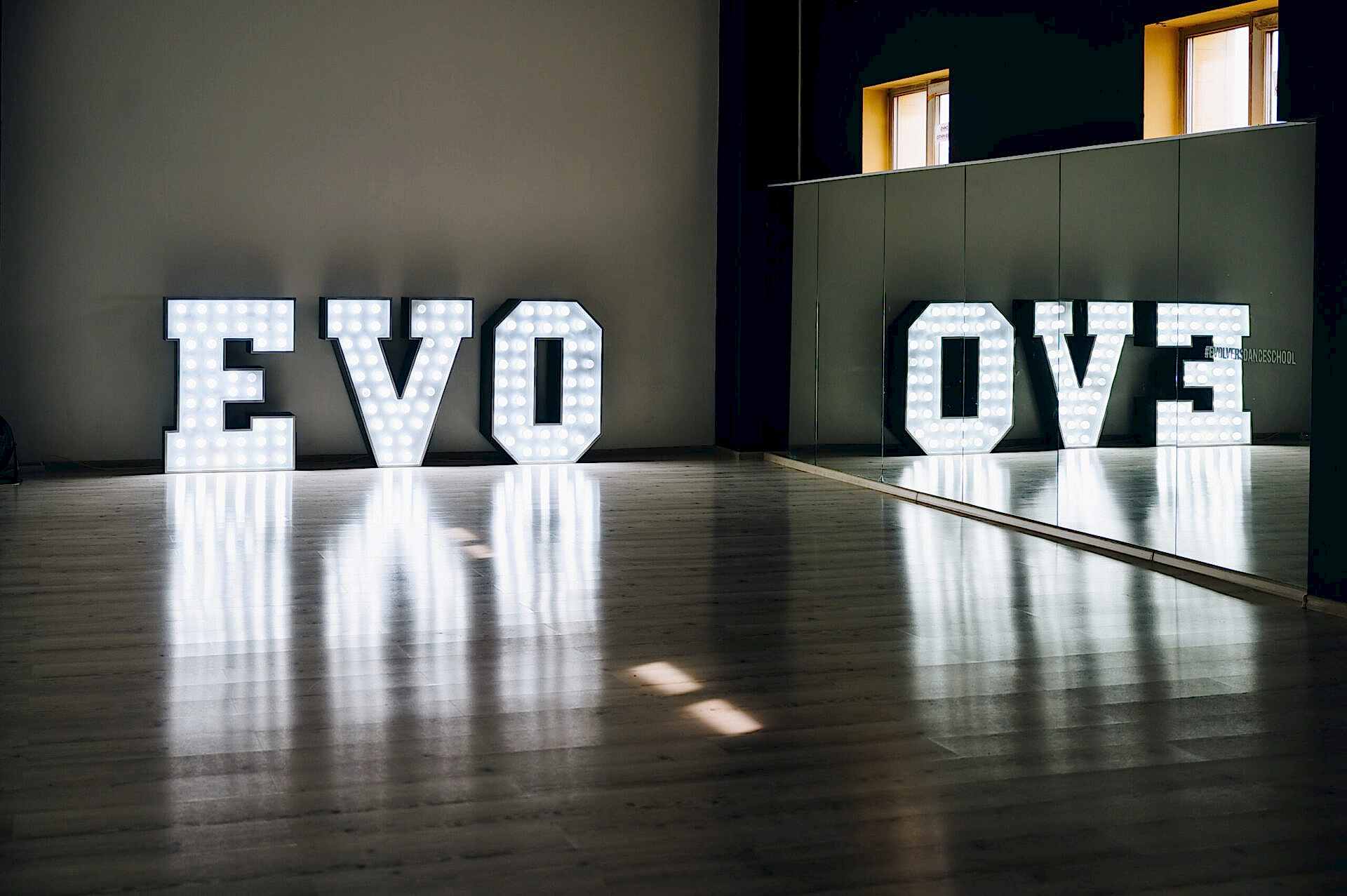 Свечение букв EVO в помещении с отражением в зеркале на деревянном полу, создающее атмосферу.